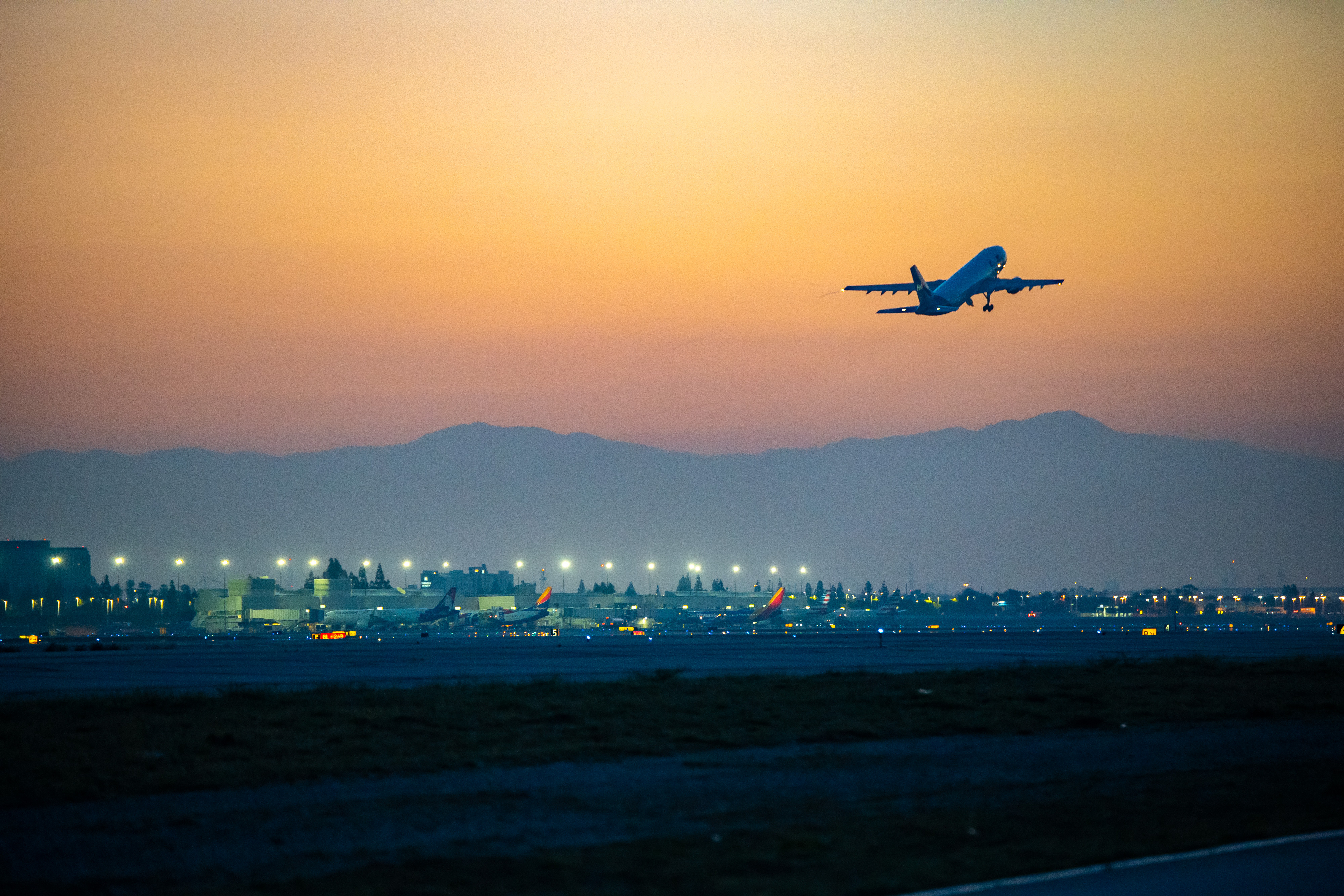 Silhouette of airplane taking off at sunset over city skyline with mountains in background.