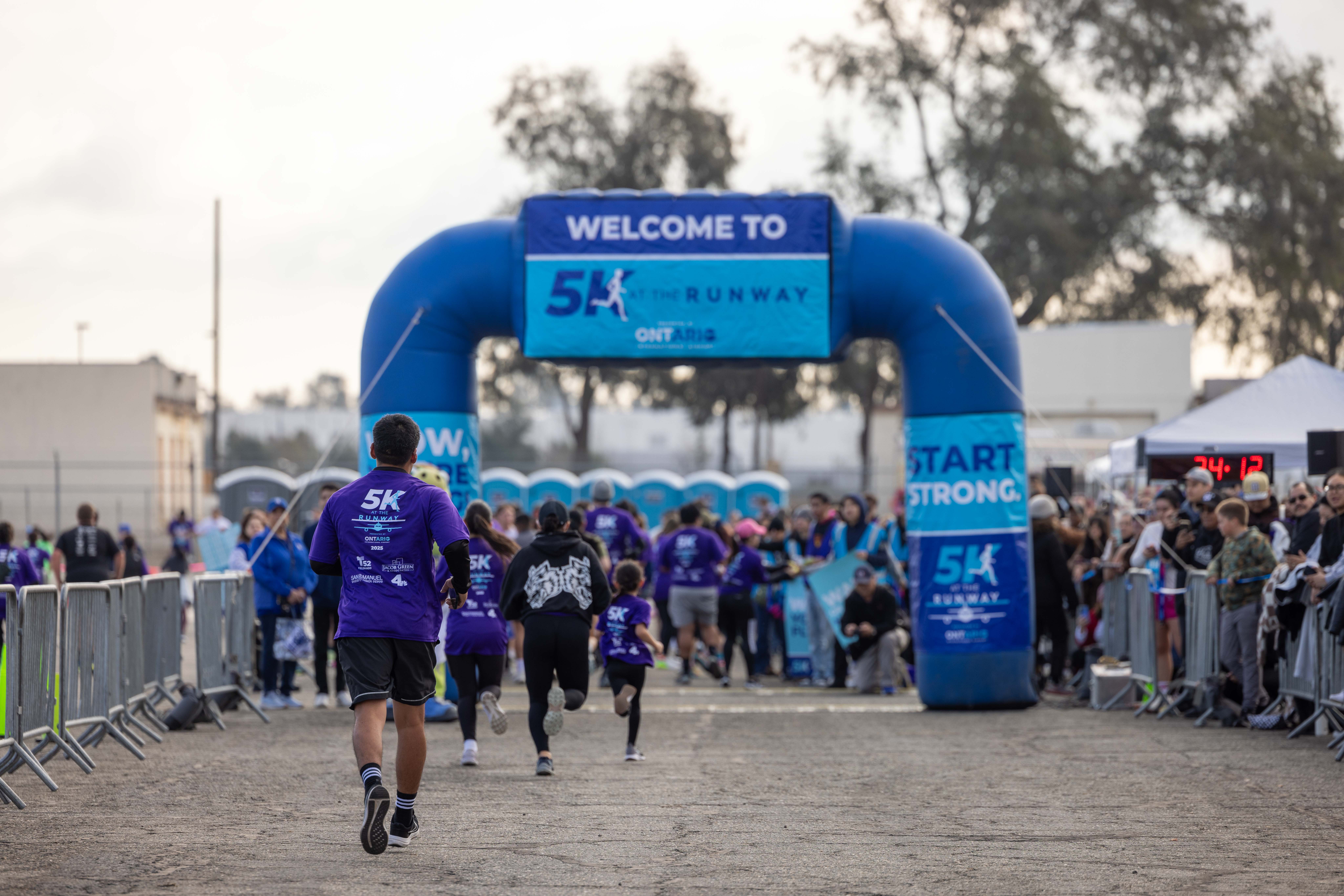 The backs of many runners are seen as they're about to cross under the inflatable blue finish line marker as supporters stand on the sidelines.
