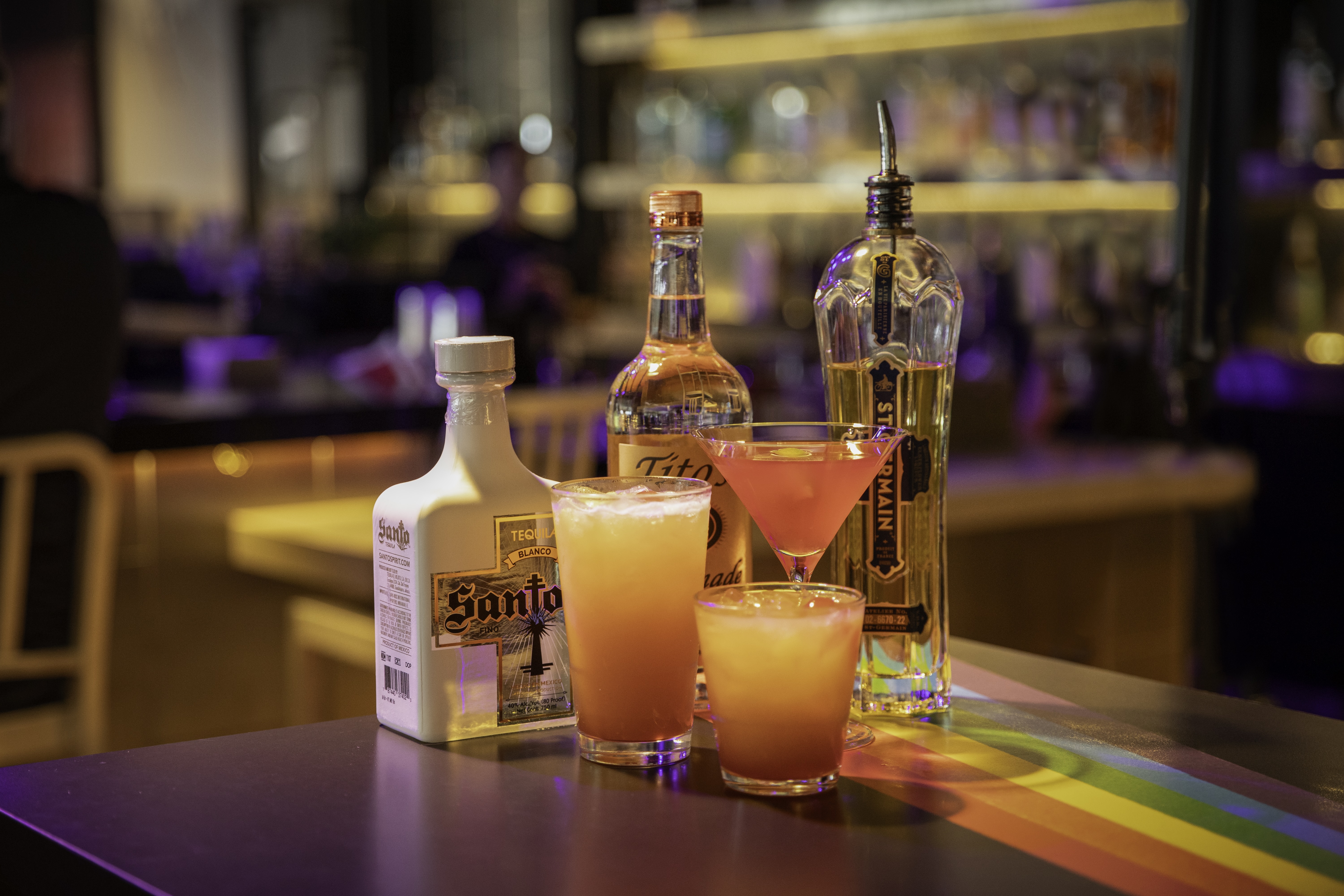Two cocktail glasses with orange drinks next to a bottle in a dimly lit bar setting.