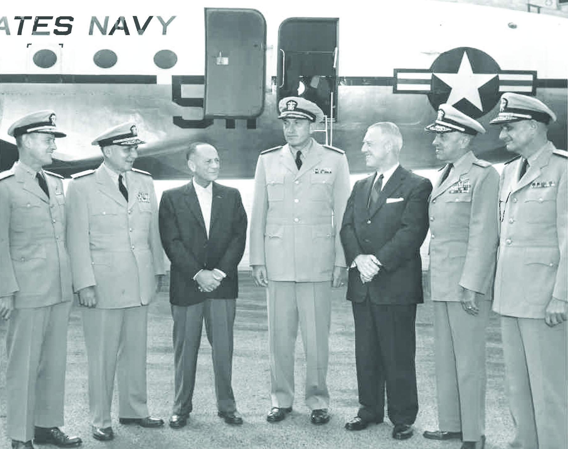 In this black and white photo, a group of seven men, with five in military uniforms and two in suits, stand in front of a Navy plane at ONT.