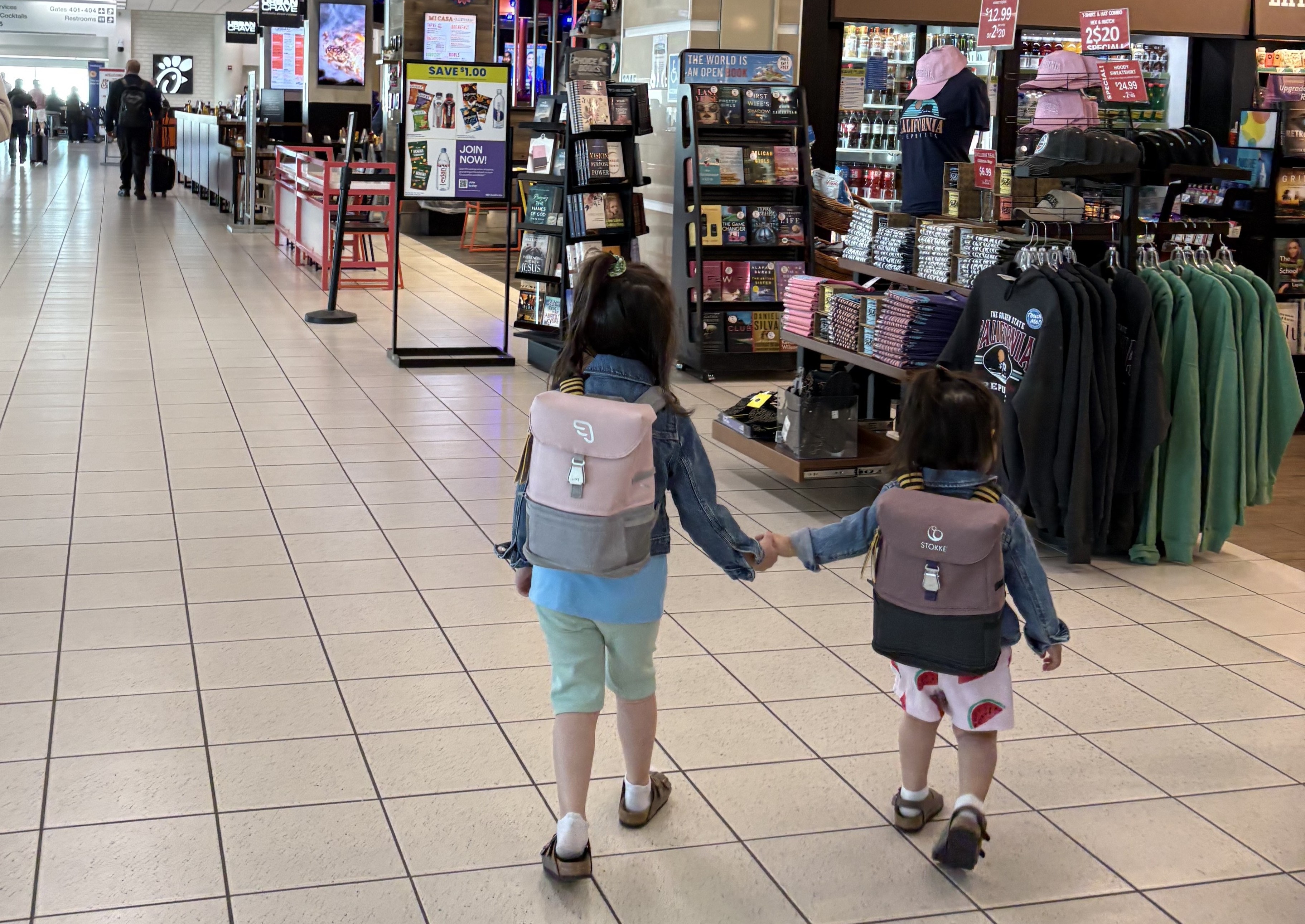 Two children walking hand-in-hand through a terminal with retail stores on both sides.