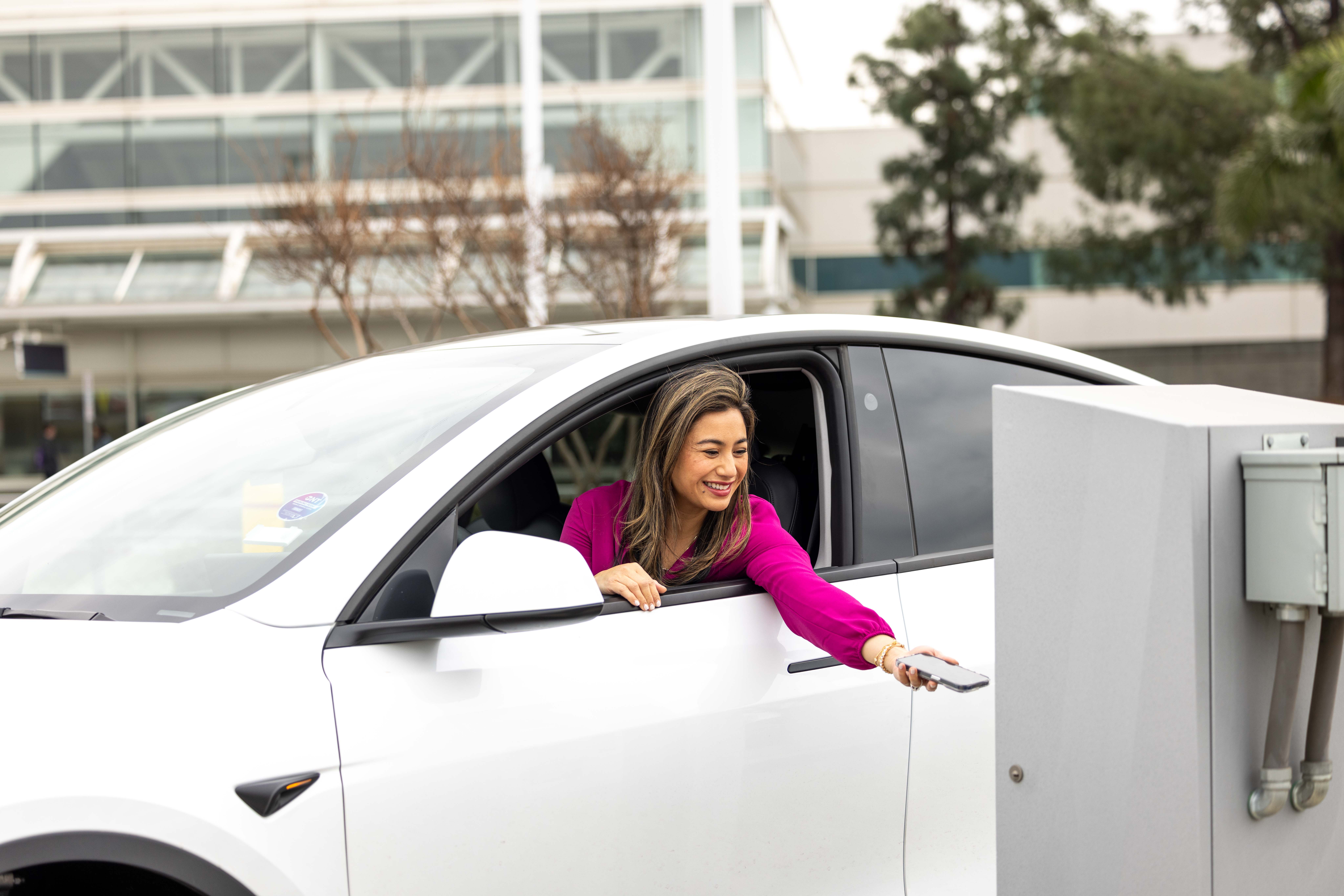A passenger reaches out the window of her white car, holding her smartphone in her hand to scan a QR code at the parking lot entry machine.