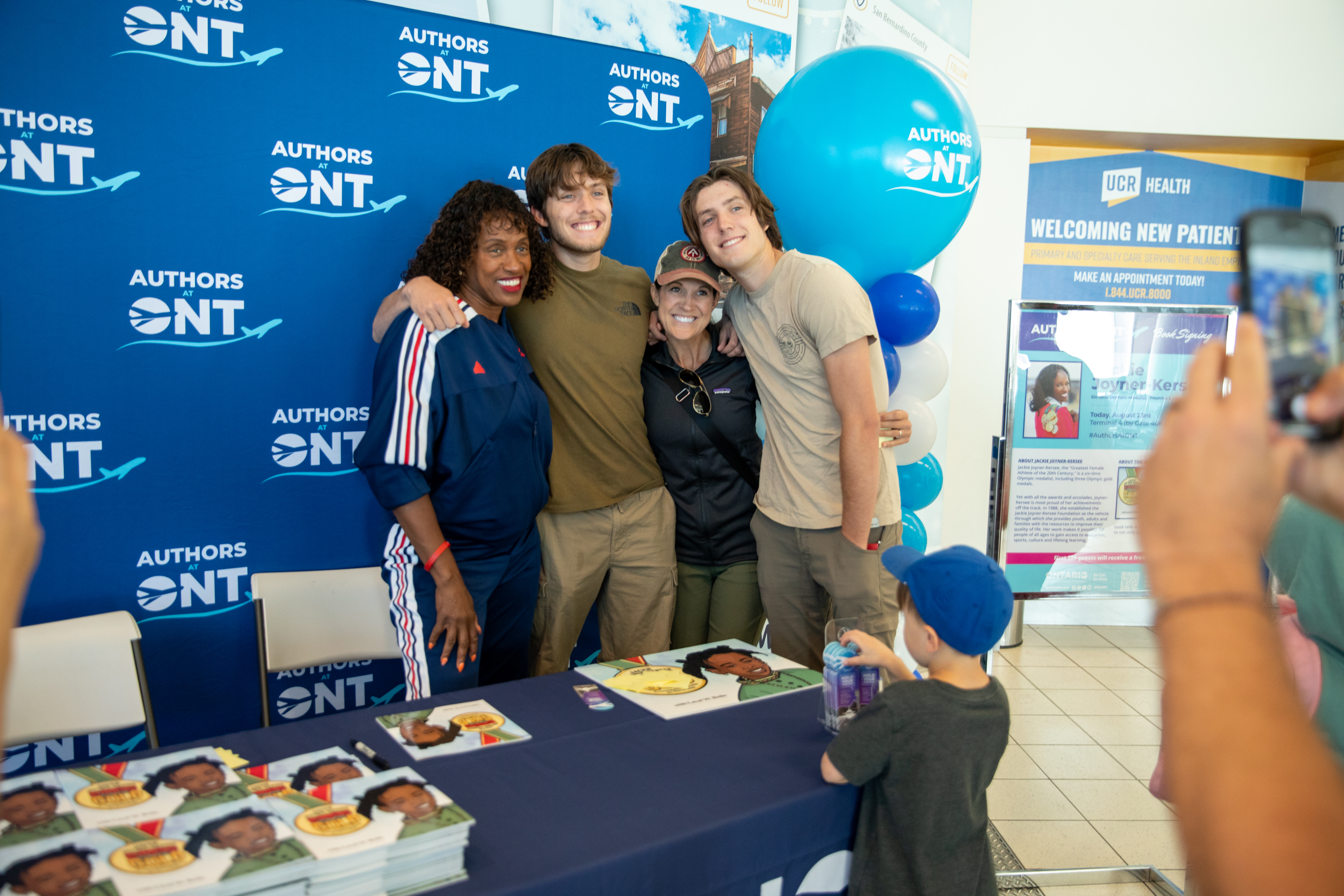 Olympian Jackie Joyner-Kersee poses for a photo with three passengers in front of a table with her children's book on display.