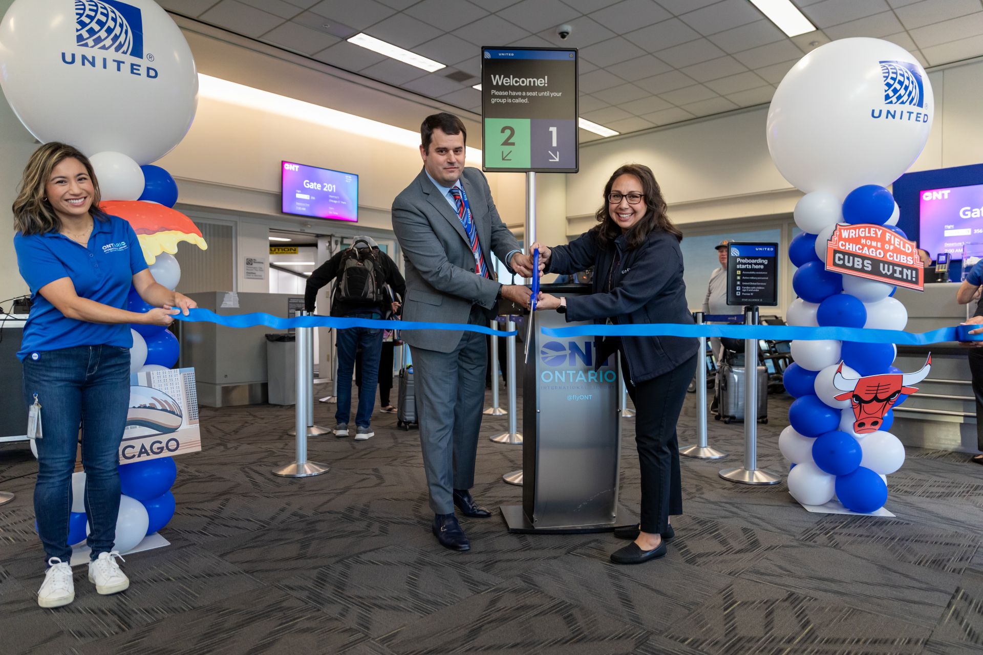 Two people use large scissors to cut a blue ribbon with big white balloons that say United on each side.