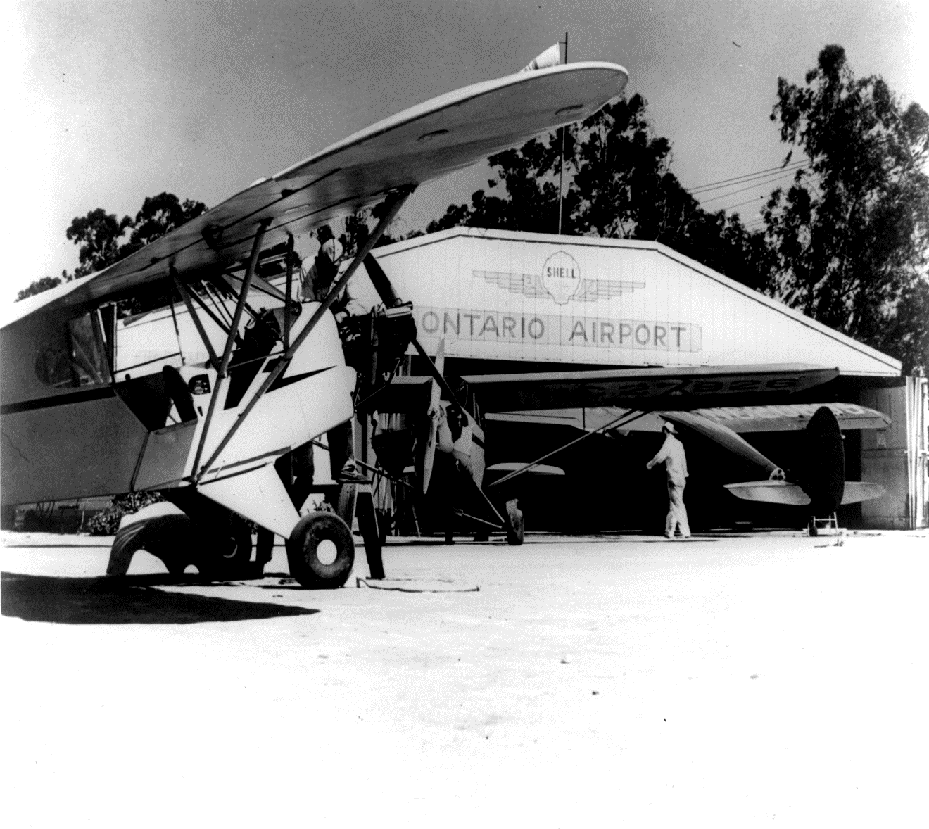 Black and white photo shows a hangar that says Ontario Airport, with a man walking in front and prop planes parked nearby.