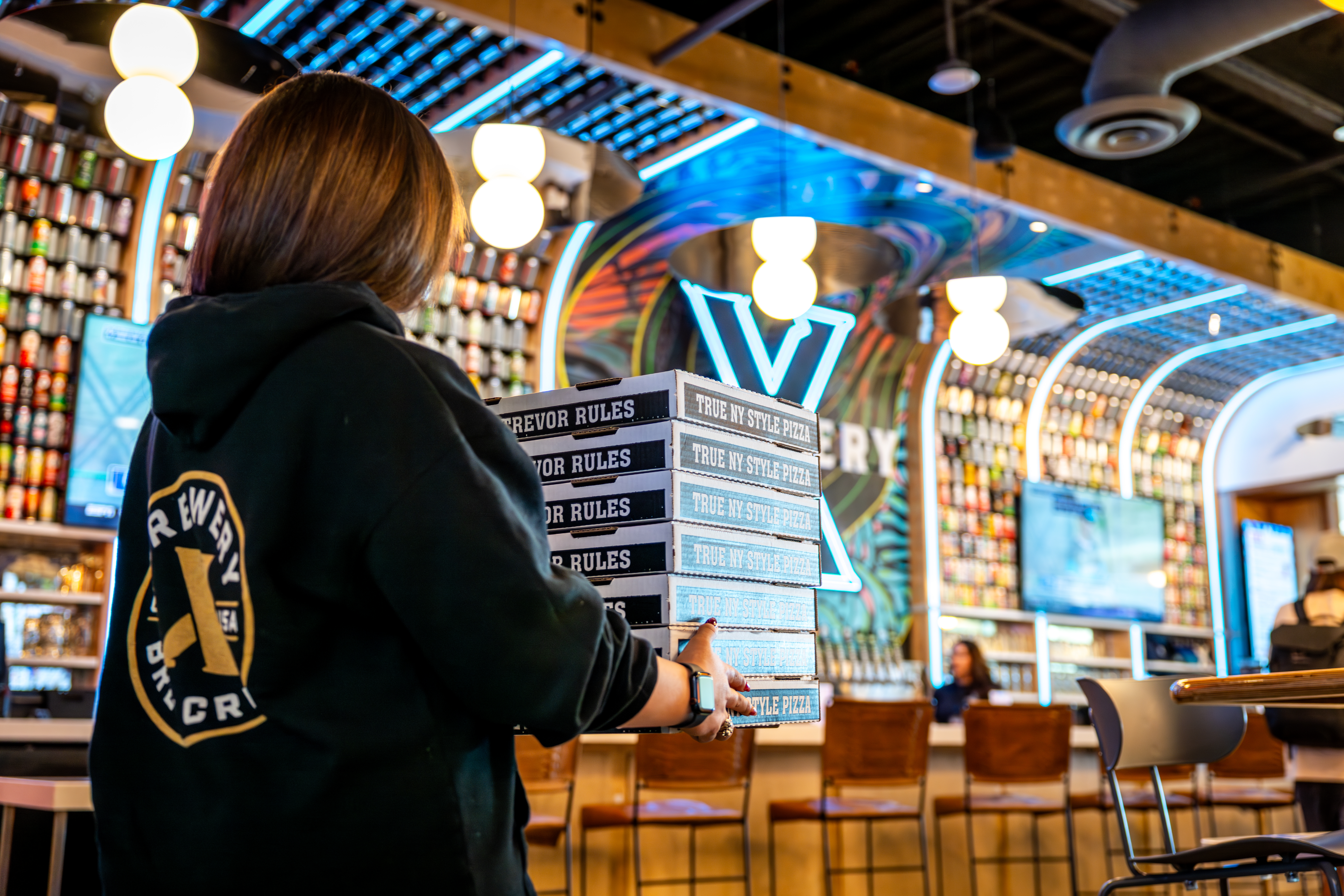 Brewery X employee delivering a stack of pizza boxes across the restaurant with the bar in the background