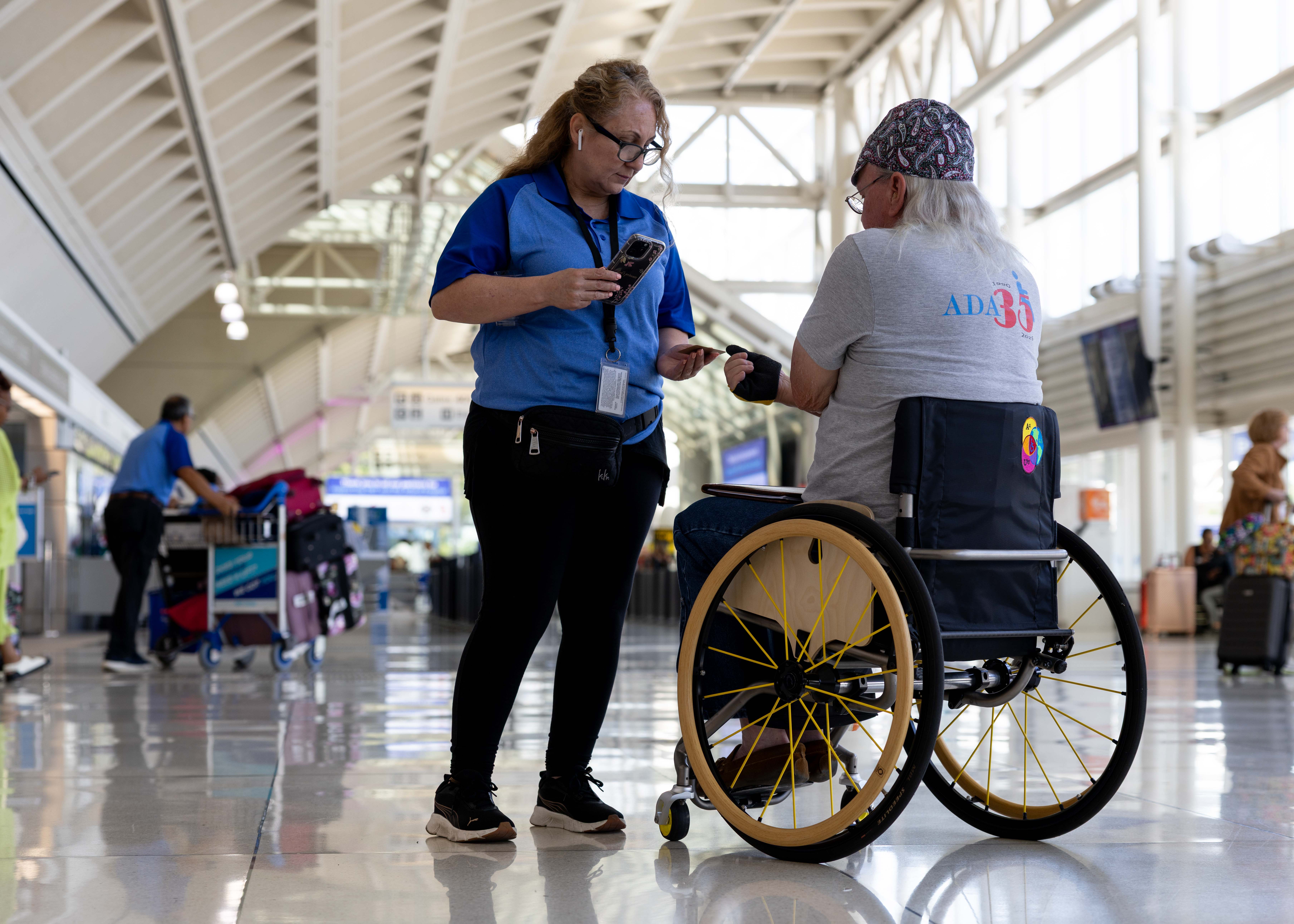 A staff member in a blue shirt scans the boarding pass of a passenger in a wheelchair on the first floor of an ONT terminal.