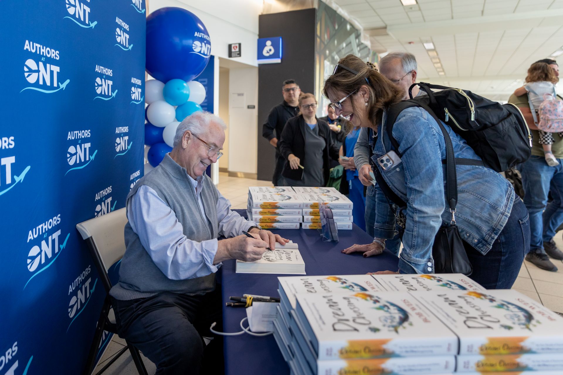 Author Bob Weis signs a book for a smiling passenger with a stack of his books on a table in the terminal.