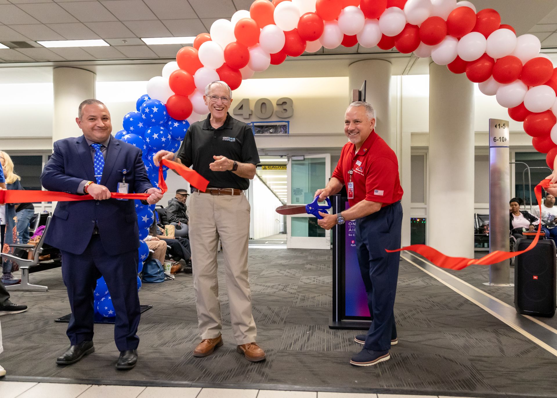 Three men cut red ribbon while smiling with a red and white balloon arch behind them in an ONT terminal.