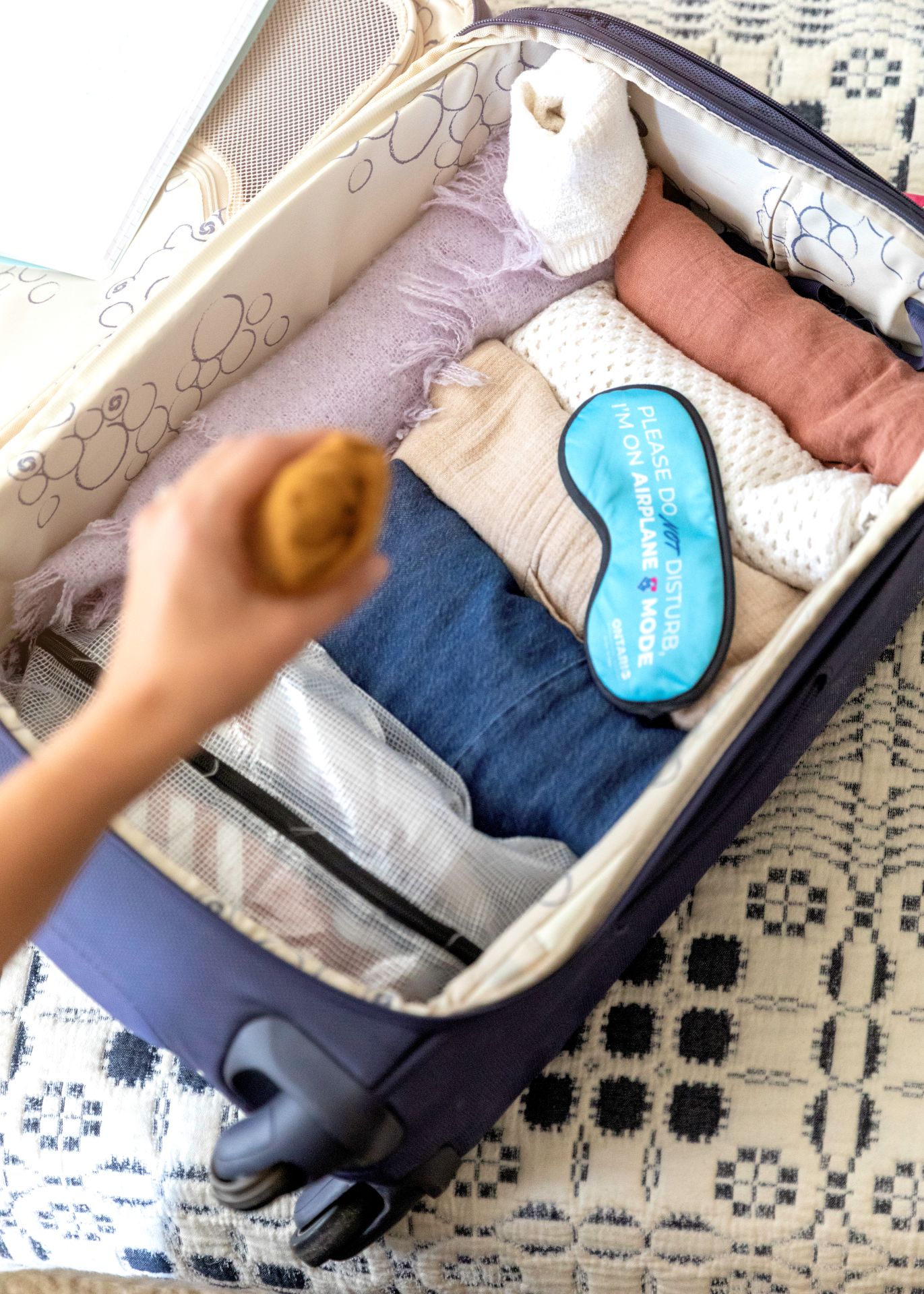 A hand is throwing a scarf into a carry-on suitcase that has rolled clothing with an ONT sleep mask on top.