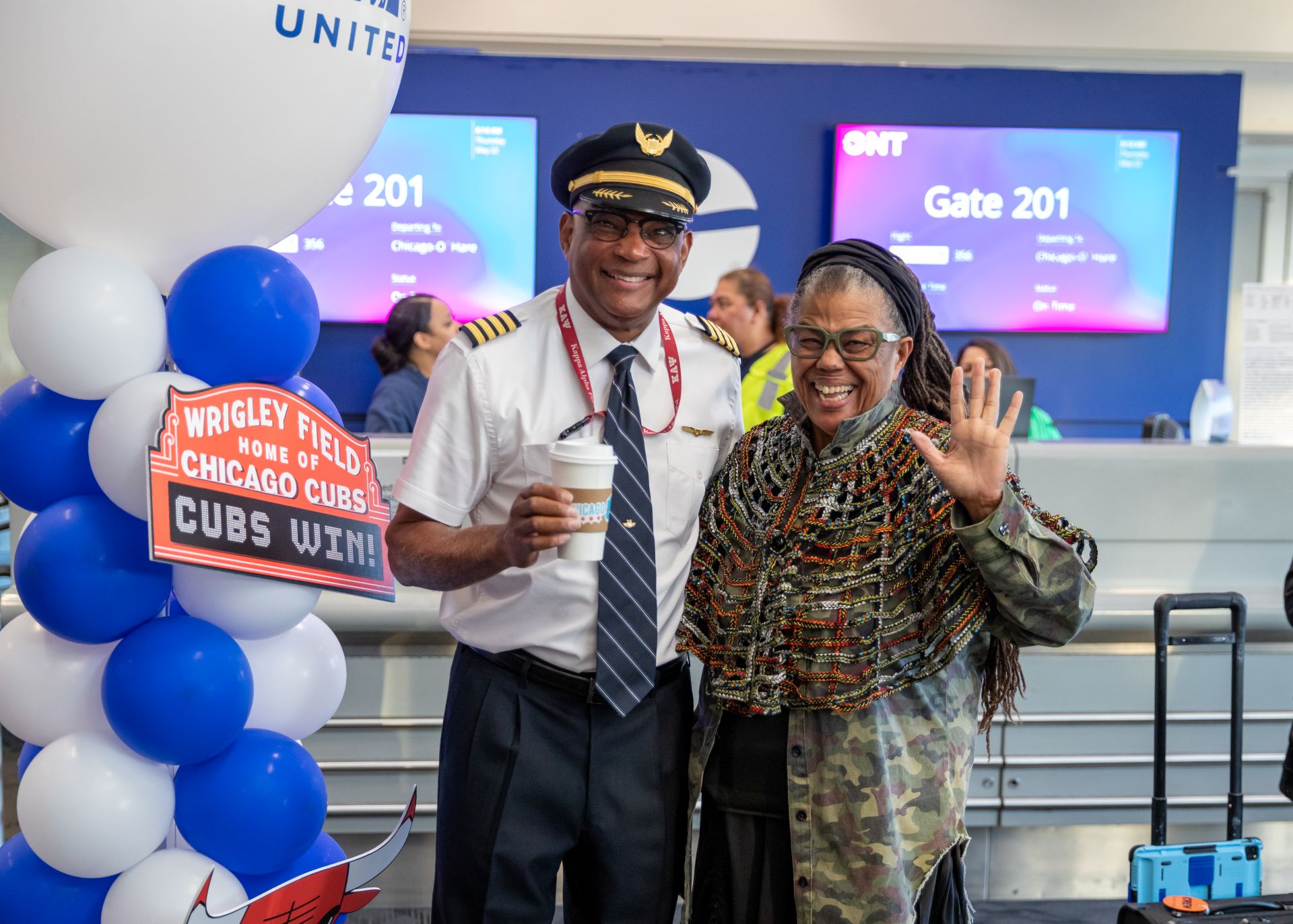 A pilot stands with a passenger waving at the camera. He's holding a Chicago coffee cub with a Wrigley Field sign behind them.
