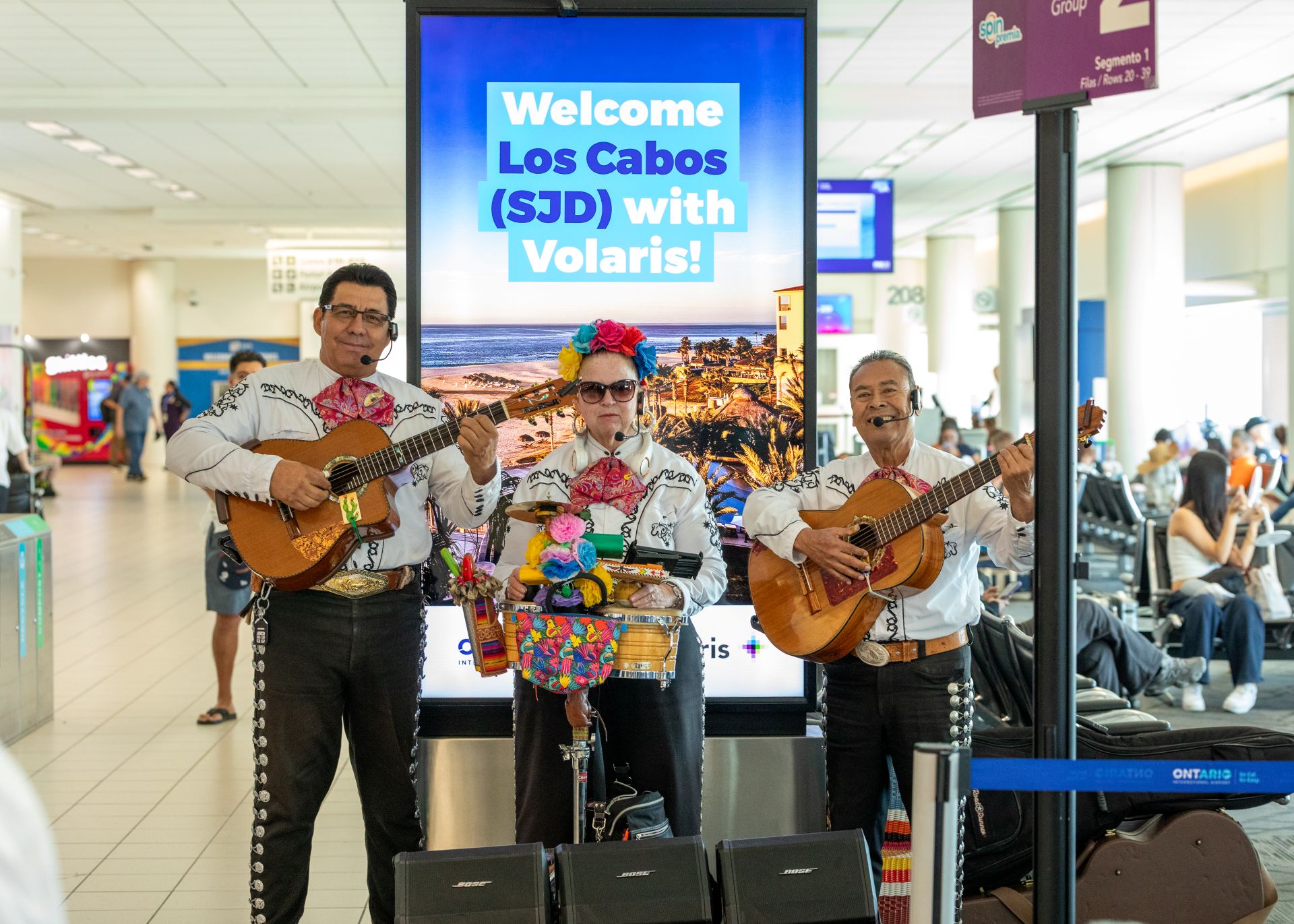 Three musicians perform in traditional Mexican clothing in front of a sign that says Welcome Los Cabos at ONT.