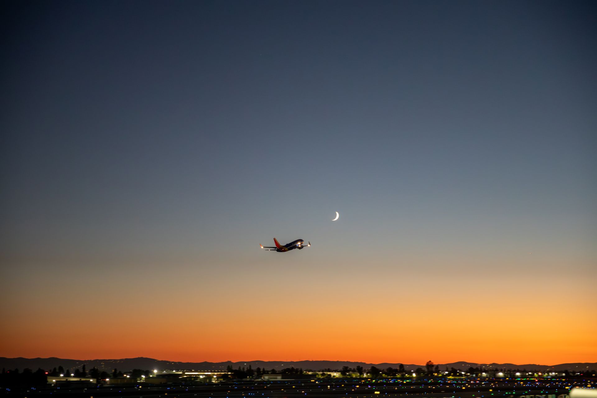 A plane is shown taking off at dusk with a narrow moon in the sky.