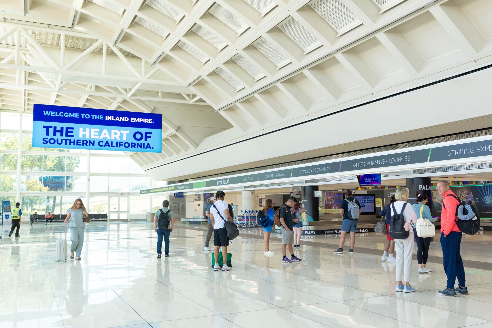 Passengers are seen in the baggage claim area with a sign that says Welcome to the Inland Empire, the Heart of Southern California.