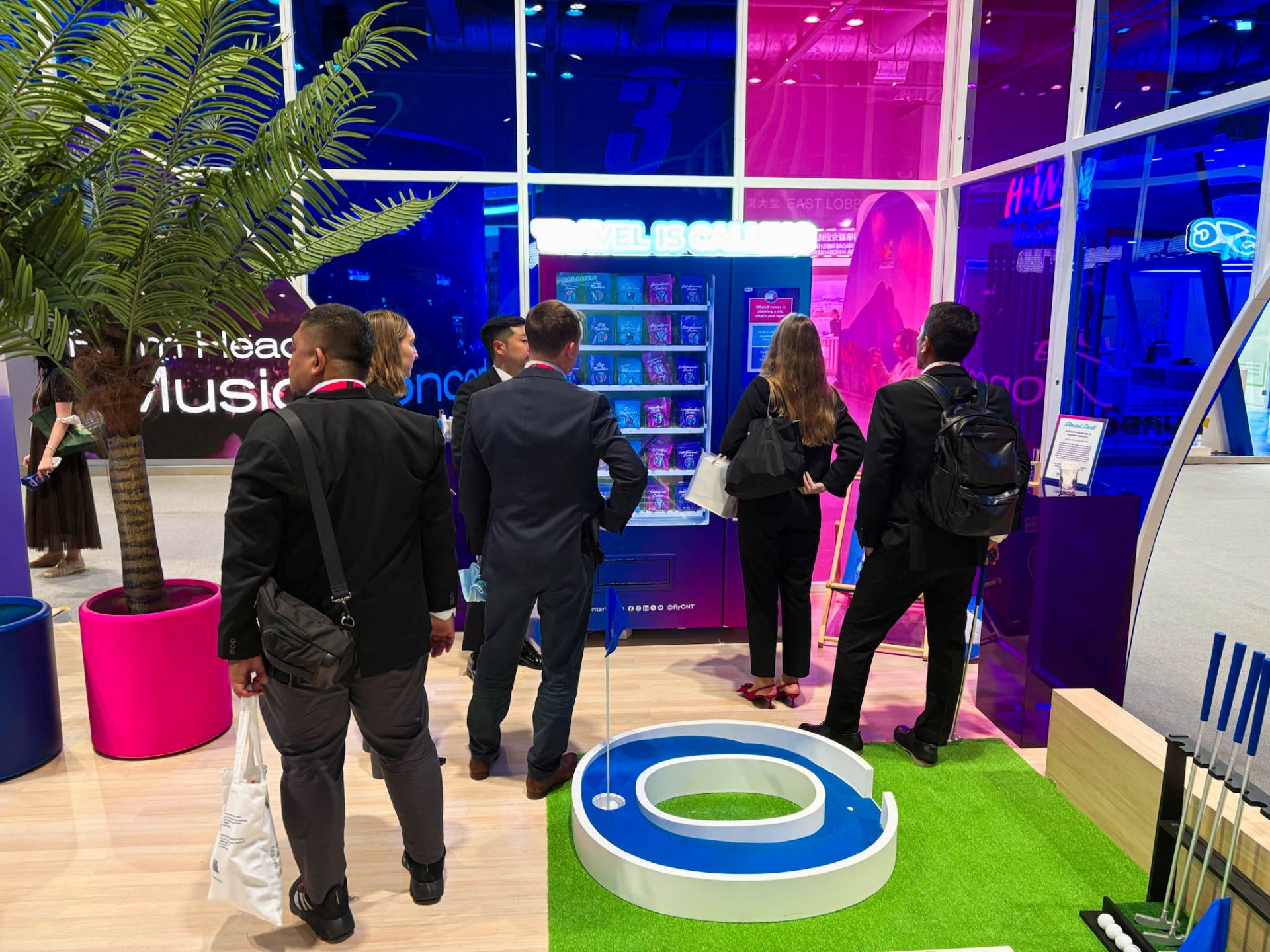 A group of people in suits stand inside a brightly colored ONT booth with a putting green, plants and an ONT-branded vending machine.