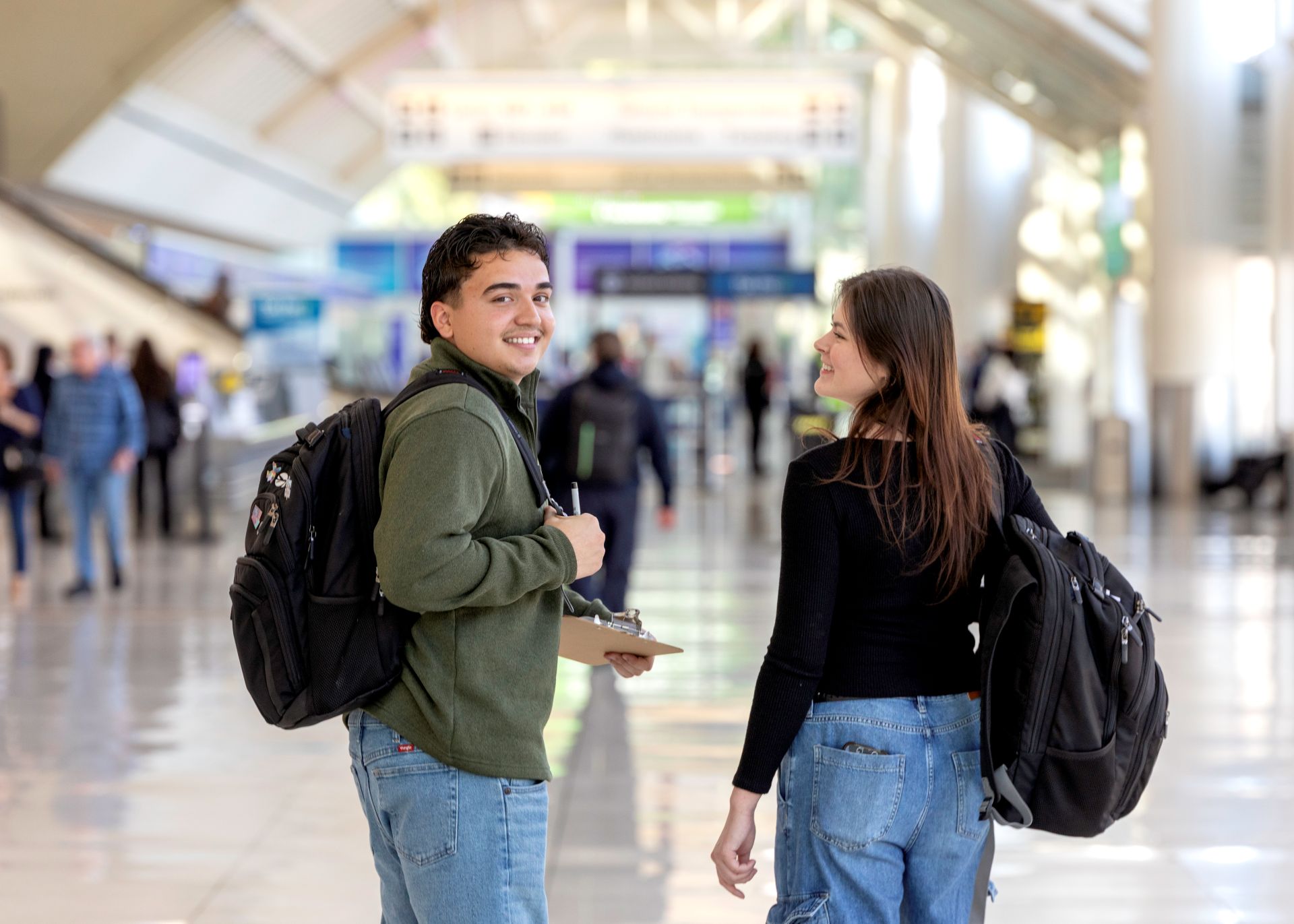 A young man looks at the camera as he walks beside a young woman through an ONT terminal with both wearing black backpacks.
