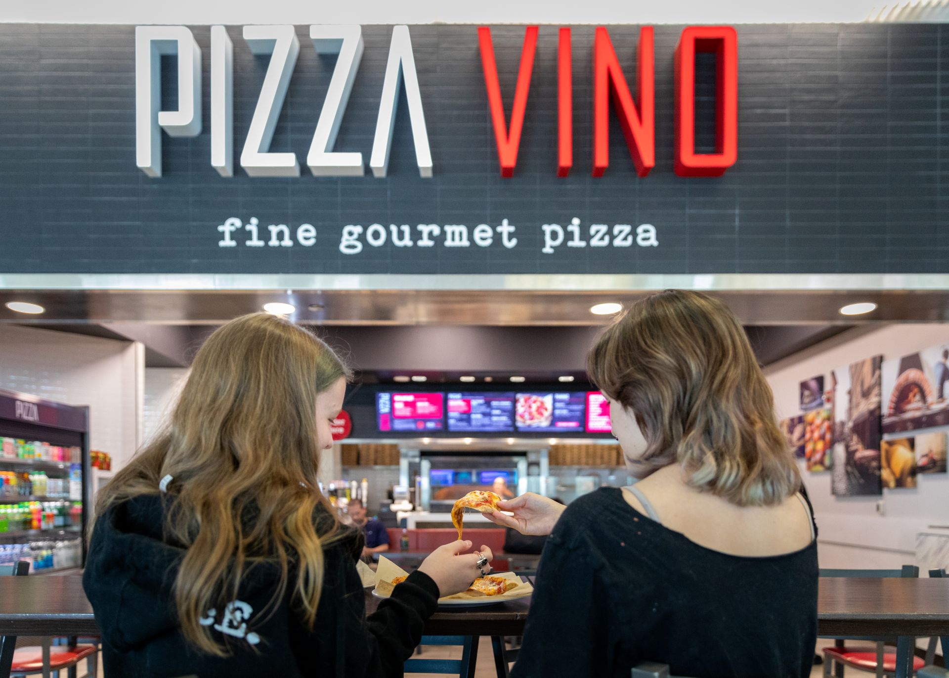 Two young women with their backs to the camera sit in front of Pizza Vino eating pizza.