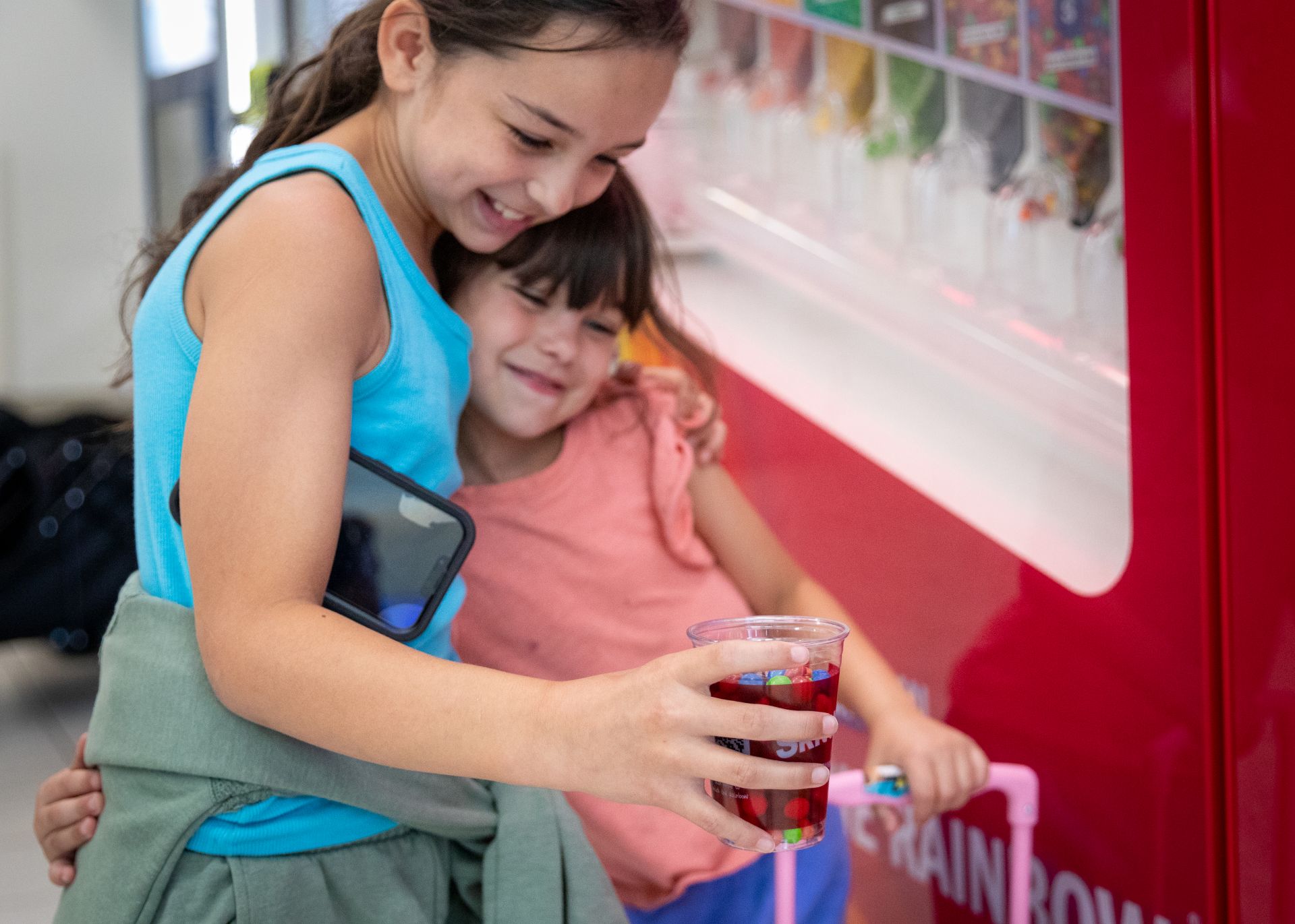 Two young girls smile and hug while holding a clear plastic cup full of Skittles.