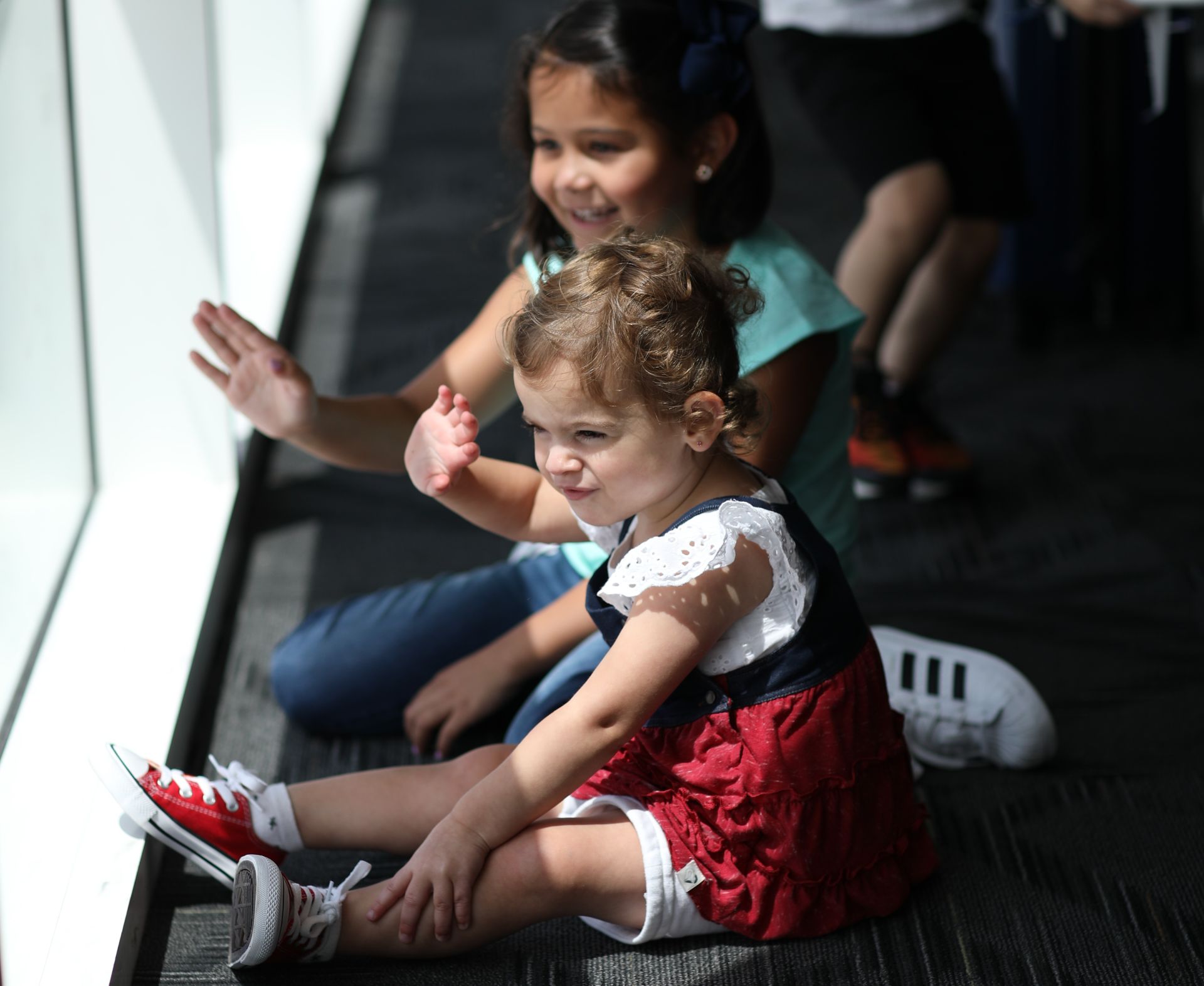A toddler and a young girl look at the airport window and wave at planes as they sit on the ground smiling.