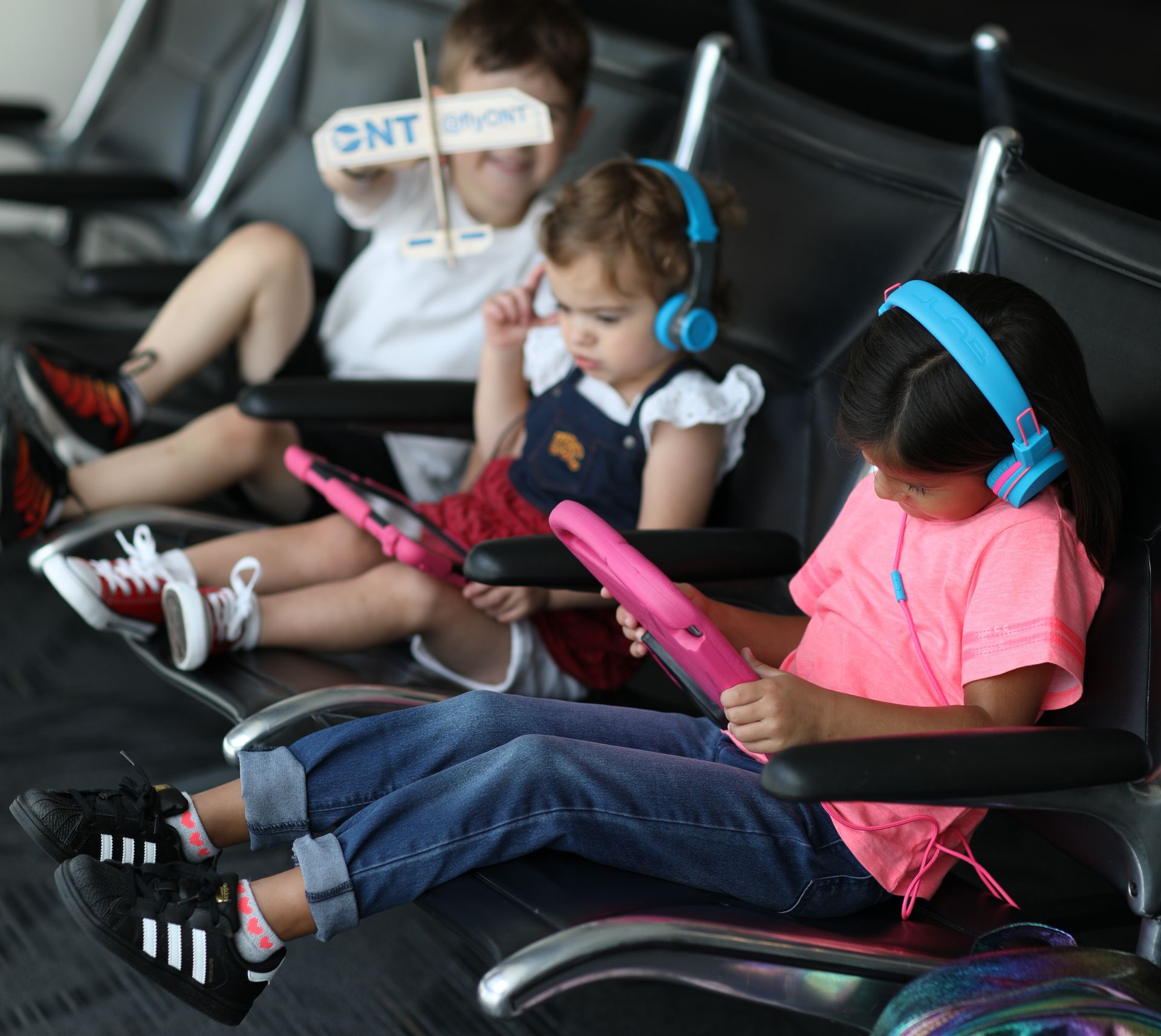 Three kids sit in seats at an airport, with two girls looking at tablets while a boy plays with an ONT wooden plane.