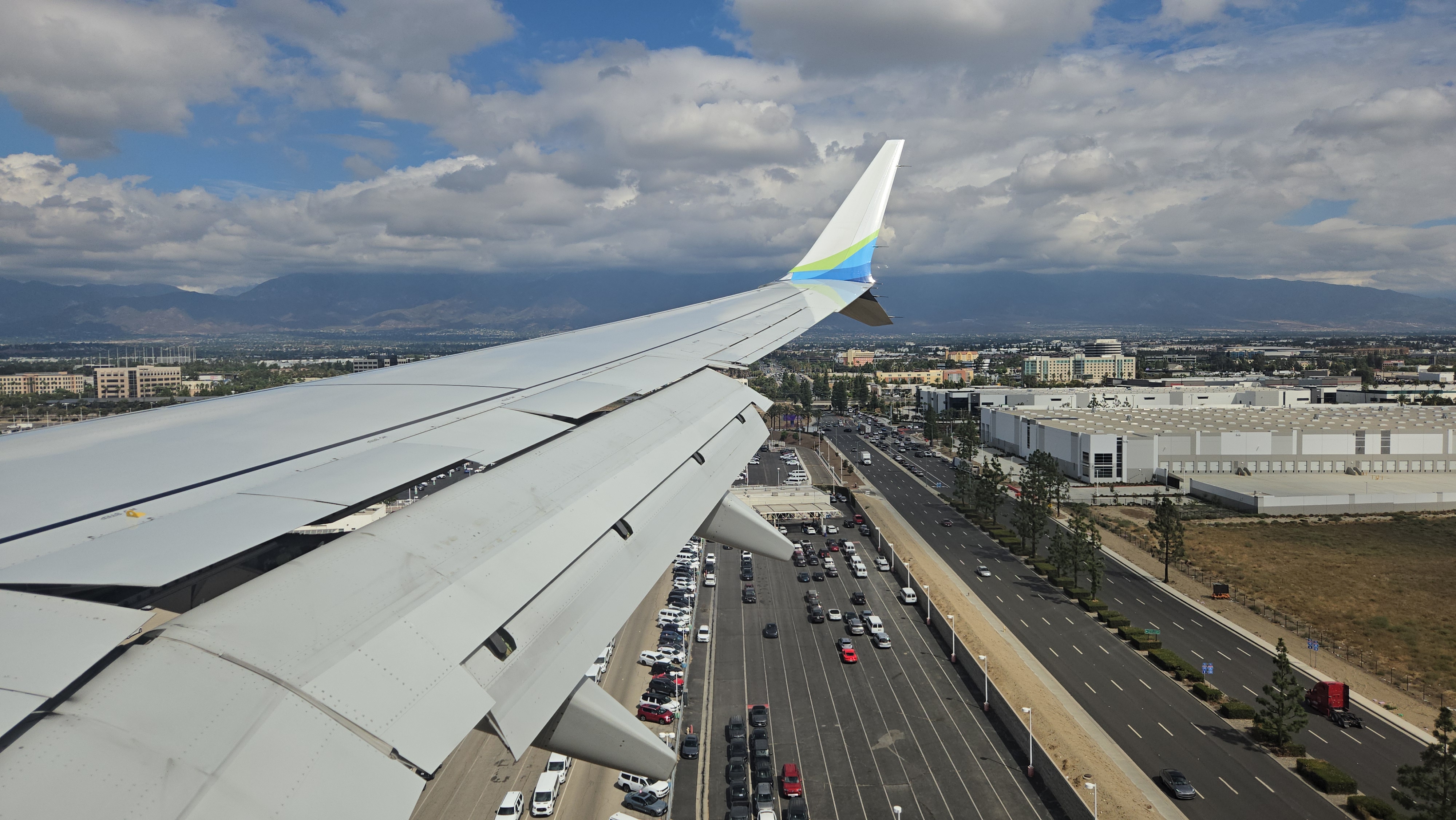 The wing of a plane is seen flying over a busy freeway with mountains in the background.