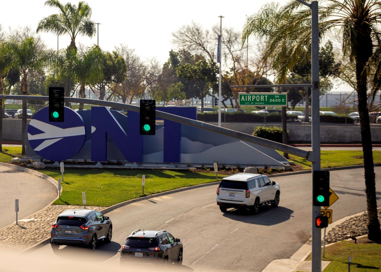 Cars drive under the Airport Drive road sign to enter ONT past a blue and white ONT monument sign.