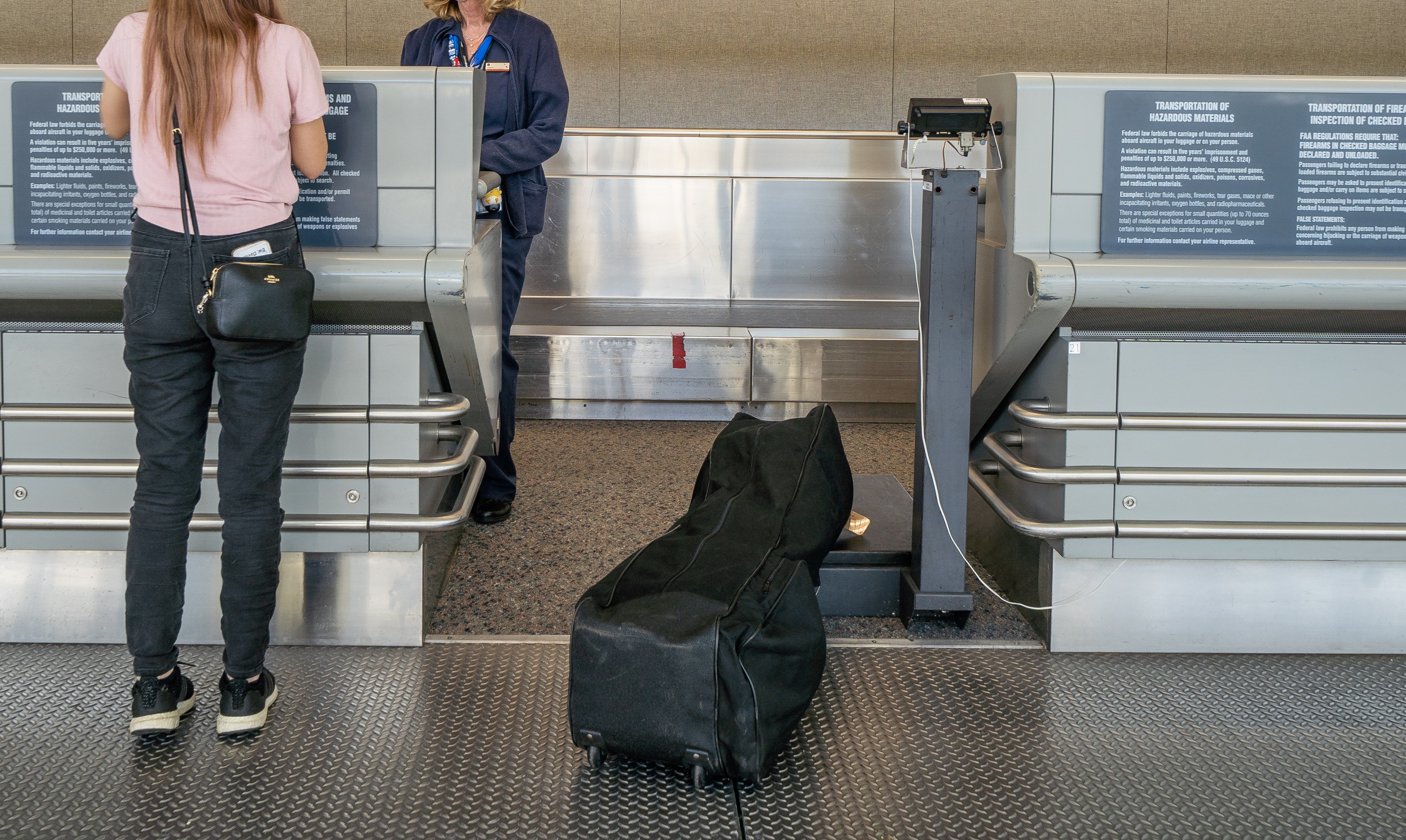 A black soft guitar case is seen sitting on the scale at an airline ticket counter with a woman standing nearby talking to an agent.