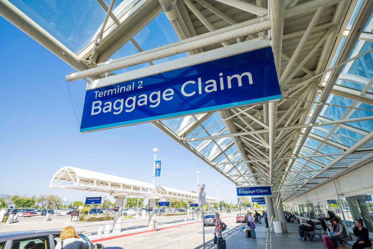 A blue sign with white writing says Baggage Claim over a bustling sidewalk of passengers with a roadway nearby.