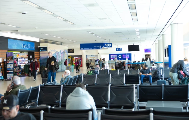 Passengers seated and walking through an airport gate area with visible signs and terminal shops in the background.