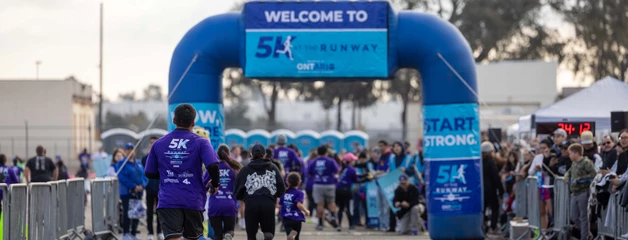 The backs of many runners are seen as they're about to cross under the inflatable blue finish line marker as supporters stand on the sidelines.