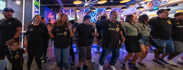 A line of people with arms over each other's shoulders dance in celebration of LAFC inside Brewery X at ONT.