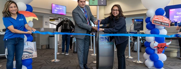 Two people use large scissors to cut a blue ribbon with big white balloons that say United on each side.