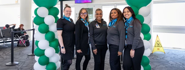 Five members of the Frontier flight crew pose near green Frontier ballons towers with a sign promoting the new flight to Portland, Oregon behind them.