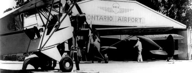 Black and white photo shows a hangar that says Ontario Airport, with a man walking in front and prop planes parked nearby.