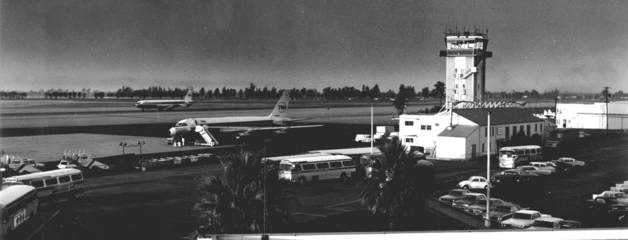 An air traffic control tower, planes and parked cars are shown in a black-and-white photo.