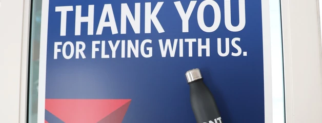 A sign says "Delta" and "Thank you for flying with us" and a black ONT water bottle is held in front of it.
