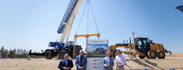 Four men stand with shovels in a dirt lot with a large crane behind them.