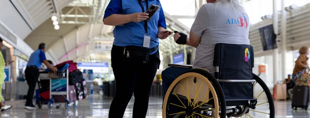 A staff member in a blue shirt scans the boarding pass of a passenger in a wheelchair on the first floor of an ONT terminal.