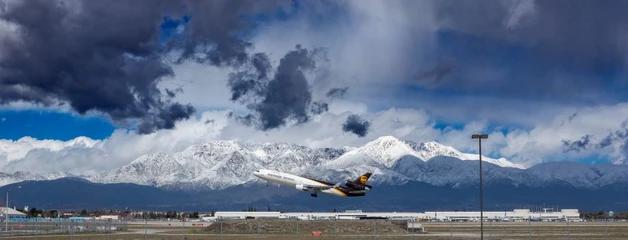 A UPS plane is shown taking off in front of the snow-capped San Gabriel Mountains.