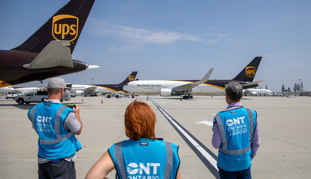 Three people in blue vests watch UPS cargo planes on tarmac