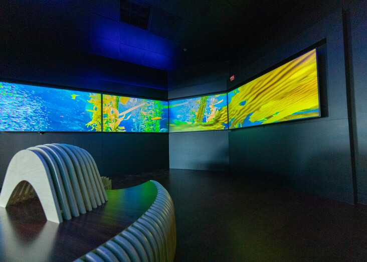 Three curved monitors displaying abstract blue, yellow and green graphics in a dimly lit room.