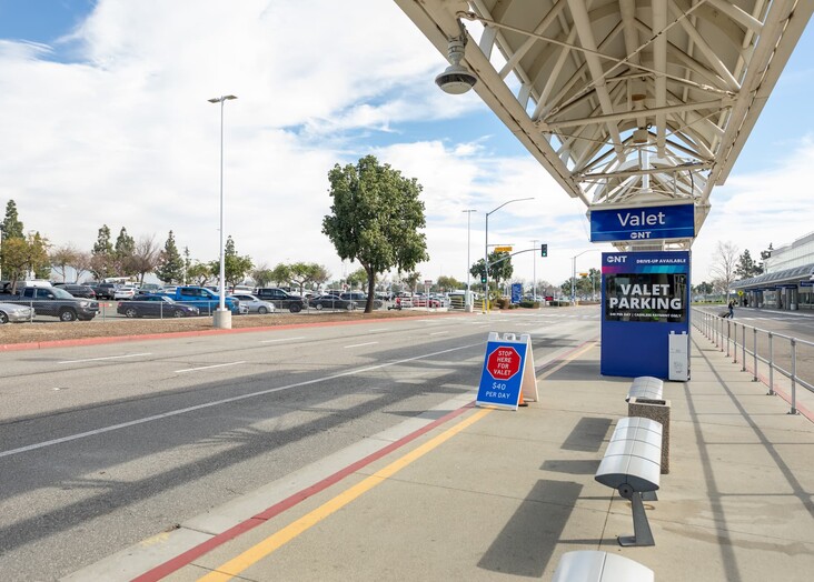 Airport terminal exterior with covered walkway, parking area, and blue sky overhead.